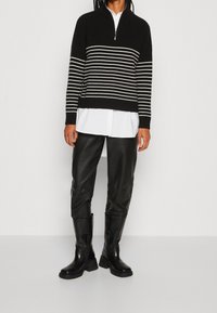 Maglione nero a maglia con strisce orizzontali bianche, colletto con zip a metà, indossato sopra una camicia bianca; abbinato a pantaloni in pelle nera e stivali chunky.