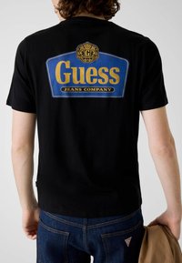 Czarny bawełniany t-shirt z dużym niebiesko-złotym logo "Guess Jeans Company" na plecach. Krótkie rękawy i standardowy krój.