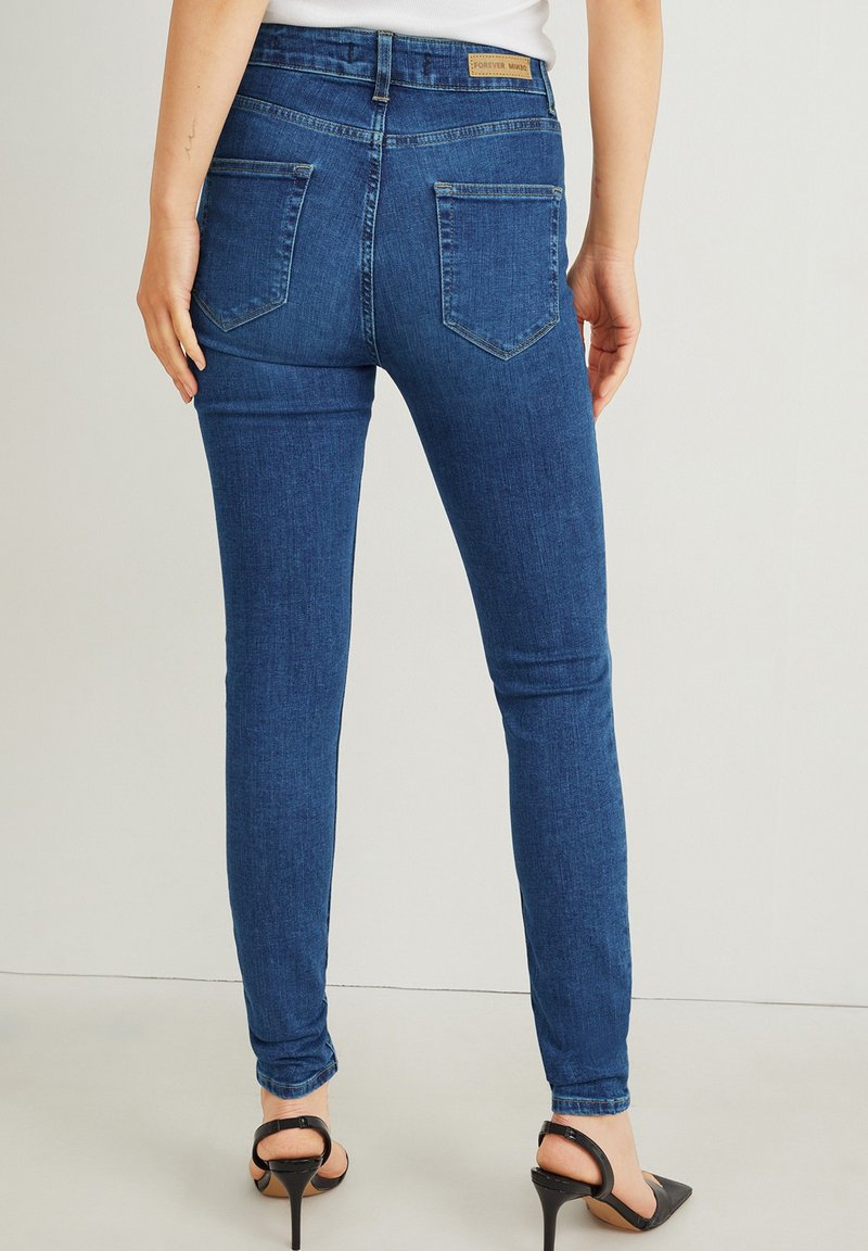 Schmale, dunkelblaue Jeans mit Gesäßtaschen, die von einer Person stehend getragen werden, kombiniert mit schwarzen, hochhackigen Slingback-Schuhen.