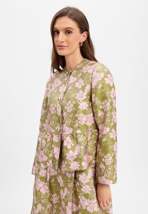 Frau trägt eine grün-pink floral gemusterte langärmelige Jacke mit Knöpfen, kombiniert mit passenden hoch taillierten Hosen, steht schräg nach links.