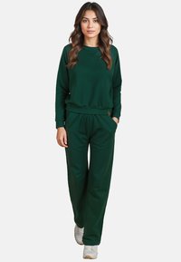Zöld pulóver és hozzáillő, puha szövetből készült nadrág, laza szabással, bordázott mandzsettákkal és funkcionális oldalsó zsebekkel.