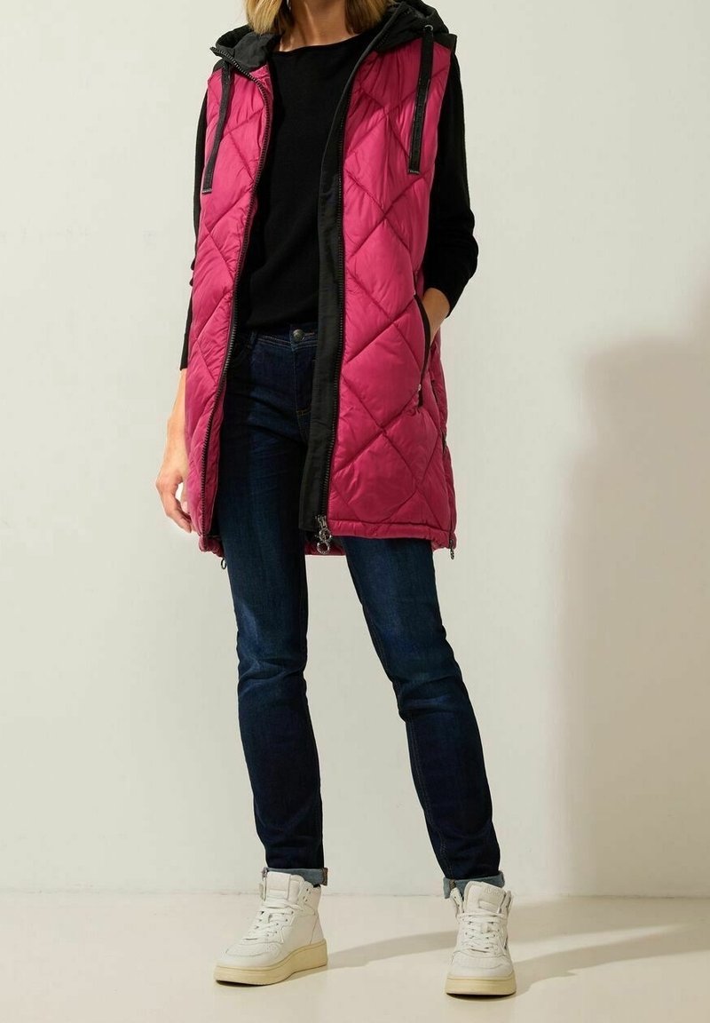 Femme portant une longue veste matelassée rose avec capuche noire, un t-shirt à manches longues noir, un jean sombre et des baskets montantes blanches, debout avec une main dans la poche.