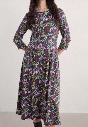 Blomstret kjole med lange ærmer, med lilla, blå og gule mønstre på en mørk baggrund. A-formet silhuet, knælang, blødt stof.