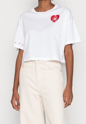 Person wearing white cropped t-shirt with red heart-shaped Converse logo on chest and small red hearts on sleeve, paired with light beige pants.