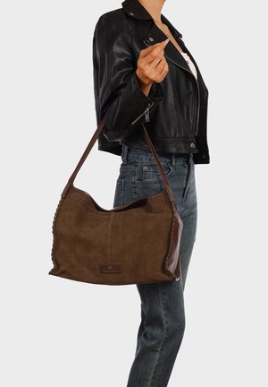 Persona con giacca di pelle nera e jeans blu che porta una grande borsa a tracolla in pelle marrone sul braccio.