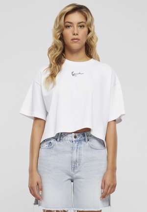 Hullámos szőke hajú nő, fehér, nagy méretű, rövidített felsőben, amin kis fekete szöveg látható, és világoskék, magas derekú farmerral.