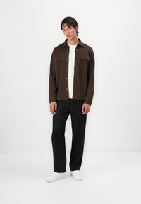 Brun button-up jacka med två bröstfickor, bärs över en vit skjorta, parat med svarta byxor och vita sneakers mot en neutral bakgrund.