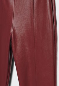 Pantaloni in pelle borgogna con una texture liscia. Presentano un design aderente e un sottile dettaglio di cuciture lungo le cuciture.