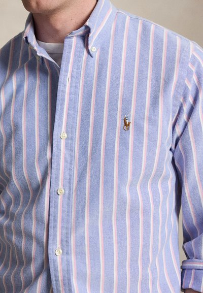 Mann iført en blå skjorte med knapper, med rosa og hvite striper og en liten brodert hestelogo på venstre bryst.