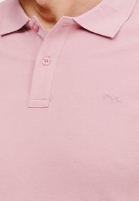 Jasnoróżowa koszulka polo wykonana z teksturowanej tkaniny, z kołnierzykiem, trzema guzikami oraz małym haftowanym logo na piersi.