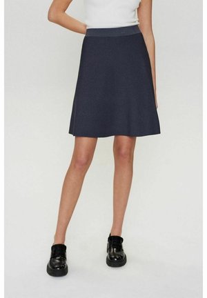 Femme portant une jupe trapèze bleu marine foncé, un haut blanc sans manches et des chaussures noires épaisses, debout devant un fond clair uni.