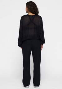 Zwart, doorzichtig gebreid sweater met lange mouwen en een gekruist ontwerp op de achterkant, gecombineerd met zwarte brede broek. De voeten zijn in sneakers.