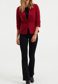 Blazer rojo con un solo botón, diseño ajustado, mangas de tres cuartos y costuras en los hombros. Combinado con pantalones acampanados negros y botines.