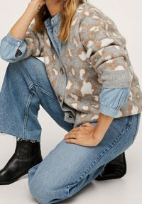 Cardigan grigio con motivo astratto marrone e bianco, abbottonato davanti, indossato sopra una camicia in denim blu e abbinato a jeans a gamba larga e stivali neri.