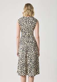 Robe sans manches en tissu beige avec des pois noirs, un corsage ajusté et une jupe évasée. Un détail de ruché au dos ajoute de la texture.