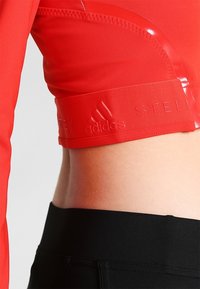 Červený sportovní cropped top s lesklými akcenty, hladkou texturou a pruhem s embosovanými logy "STELLA" a "adidas".