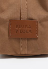Brązowa torba z materiału z wytłoczonym, sztucznym skórzanym metkiem z napisem "BIMBA Y LOLA", charakteryzująca się starannymi szwami i gładką teksturą.