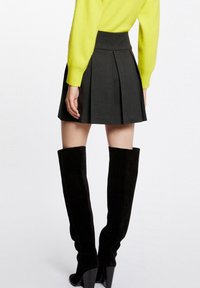 Zwarte geplisseerde rok met een hoge tailleband, gecombineerd met zwarte suède laarzen tot op de dijen en een felgele trui. Soepele textuur, op maat gemaakte pasvorm.
