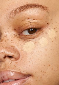 Close-up van een wang met drie kleurstalen foundation in lichte beige tinten. De huid vertoont natuurlijke sproeten en heeft een gladde, licht glanzende textuur.