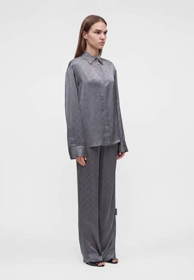 Femme debout portant une chemise ample en satin gris assortie et un pantalon large avec un motif subtil, chaussures noires à bout ouvert, fond neutre.