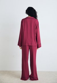 Mörkröd satin pyjamaset med långa ärmar, vida byxor och kontrasterande blå detaljer. Vertikala ränder på tyget synliga från baksidan.