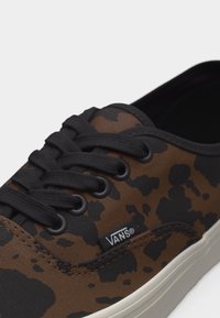 Baskets Vans camouflage marron et noir avec lacets noirs et semelle blanche, montrant l'étiquette du logo sur le côté ainsi qu'un gros plan des œillets et des coutures.