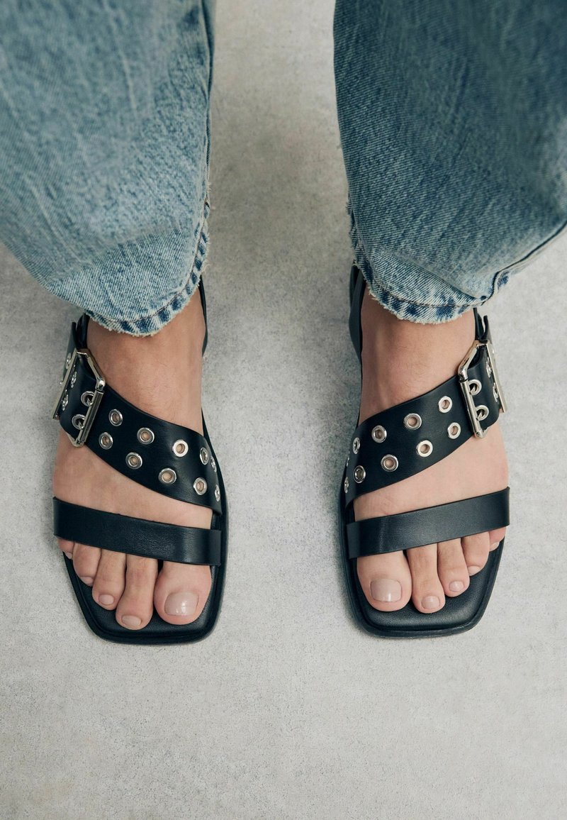Sandali in pelle nera con cinturini larghi, borchie argentate e design a punta quadrata. Abbinati a jeans azzurri chiari in stile distressed.