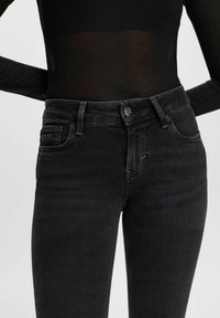 Jeans noirs taille haute slim fit portés avec un haut noir à manches longues côtelé, montrant la ceinture et les détails de la poche avant.