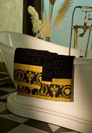 Juodas rankšluostis su auksiniais raštais ir tekstu "VERSACE". Yra sudėtingų dizainų ir tekstūrinis paviršius. Per mėlyną vonią.