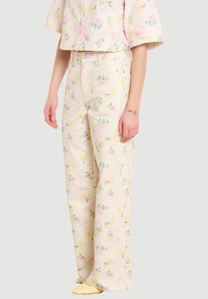 Osoba oblečená v krémových nohaviciach s vysokým pásom a zodpovedajúcej krátkej košeli s pastelovým kvetinovým vzorom, doplnená žltými špicatými topánkami a zlatým náramkom.