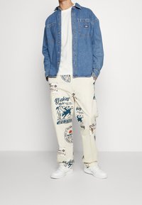 Camicia di jeans con bottoni, t-shirt bianca e pantaloni color crema con stampe grafiche blu e rosse. Sneakers bianche completano l'outfit.