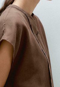 Maglietta marrone a maniche corte con collo a camicia, caratterizzata da una chiusura frontale con bottoni e un tessuto strutturato per una vestibilità comoda.