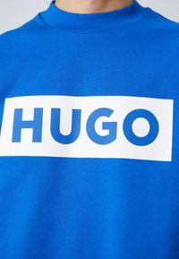 Niebieska bluza z prążkowanym dekoltem, z wyraźnym białym prostokątnym nadrukiem z napisem "HUGO" w pogrubionych niebieskich literach.