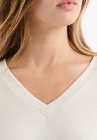 Beżowy top z dekoltem w serek o miękkiej fakturze, z czystym szwem przy linii szyi i subtelnymi tonami skóry widocznymi przy obojczyku.