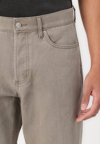 Beige denim jeans with a straight cut, single front pocket, metal button closure, and visible stitching detail on the seams.