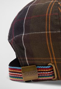 Καφέ καπέλο με καρό σχέδιο και αντίθετες πορτοκαλί, λευκές και κόκκινες γραμμές. Ρυθμιζόμενο πολύχρωμο λουράκι με ατσάλινο λογότυπο.