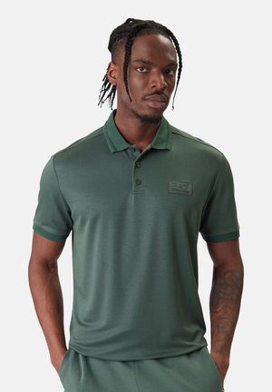 Hombre con el pelo trenzado lleva una camisa polo EA7 verde oscuro y pantalones a juego, de pie con las manos en los bolsillos frente a un fondo liso.