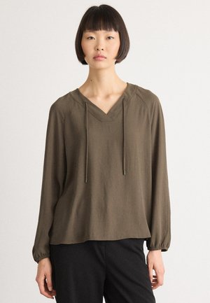 Giovane donna con capelli corti e scuri che indossa un ampio top verde oliva con maniche lunghe, scollatura a V e lacci, abbinato a pantaloni neri.