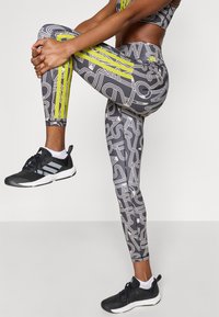 Κολάν σε μαύρο, λευκό και νέον κίτρινο με γεωμετ ρικό μοτίβο· εφαρμοστή σχεδίαση· σε συνδυασμό με μαύρα αθλητικά παπούτσια με λευκές λεπτομέρειες.