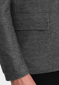 Blazer gris con un patrón de espiga, tejido texturizado y un bolsillo con solapa en el lado izquierdo del pecho.