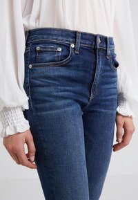Jeans de mezclilla azul oscuro con cintura alta, presentan herrajes plateados, un diseño de cinco bolsillos y detalles de costura visibles.