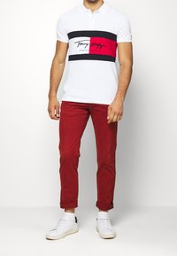 T-shirt polo blanc avec des rayures horizontales noires et rouges, accompagné d'un pantalon rouge et de baskets blanches. Texture de la peau évidente.