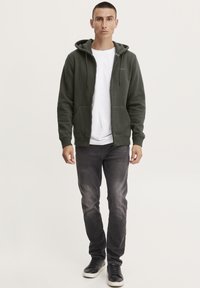 Felpa verde con zip e coulisse, tasche frontali, indossata su una maglietta bianca. Abbinata a jeans grigi scuri e sneakers nere.
