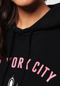Černá mikina s kapucí a růžovým grafickým nápisem "NEW YORK CITY" a bílým obrysem budovy, vybavená stahovací šňůrkou.