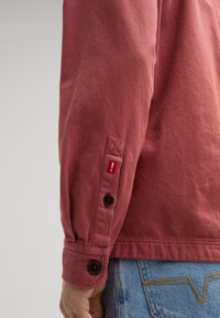 Rode langarmshirt met een textuurstof, zwarte knopen op de mouwen en een klein rood label met een uitroepteken.