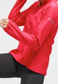 Persona vestida con una chaqueta atlética de manga larga roja brillante y leggins negros, levantando un brazo y doblando una rodilla en una posición de estiramiento.