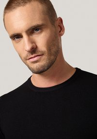 Czarny sweter dzianinowy z okrągłym dekoltem, dopasowany krój i fakturowane detale, noszony przez osobę z krótkimi włosami.