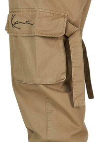 Beige cargobyxor i bomull med sidoficka, har ett knytband i midjan och en svart broderad logotyp på fickan.