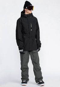 Veste noire imperméable à col haut, poche zippée à l'avant, associée à un pantalon gris. Accessoires : gants et bonnet. Lunettes de neige sur le front.