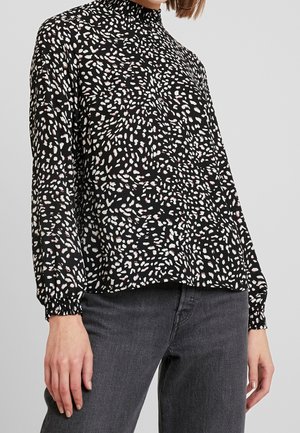 Femme portant un chemisier noir à manches longues avec des taches abstraites blanches et roses, associé à un jean gris foncé, sur un fond clair uni.
